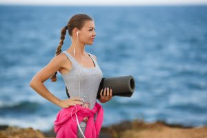 Woman standing by the ocean holding a yoga mat, wearing earphones and workout clothes.